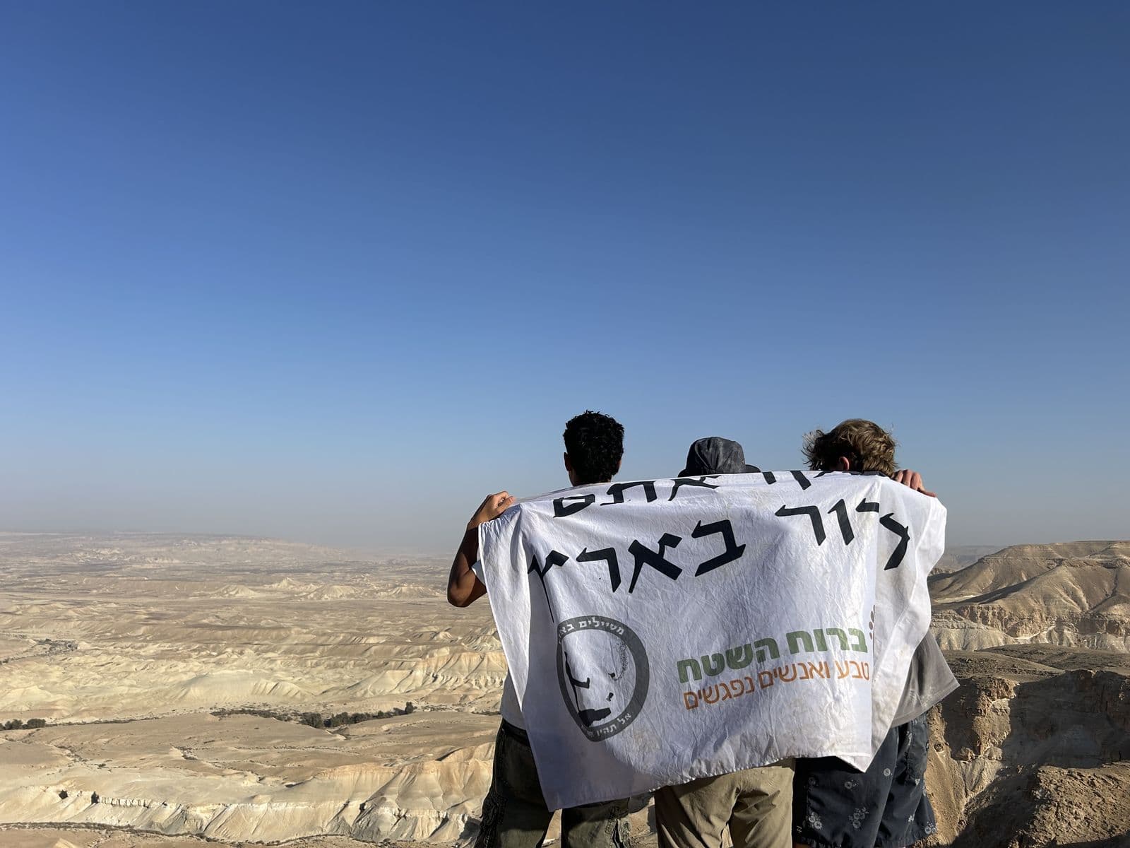 Two participants holding a B'Shetach flag against a wide desert view